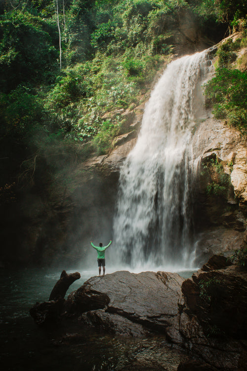 Man on a hike posing for a photo with hands up in front of a waterfall